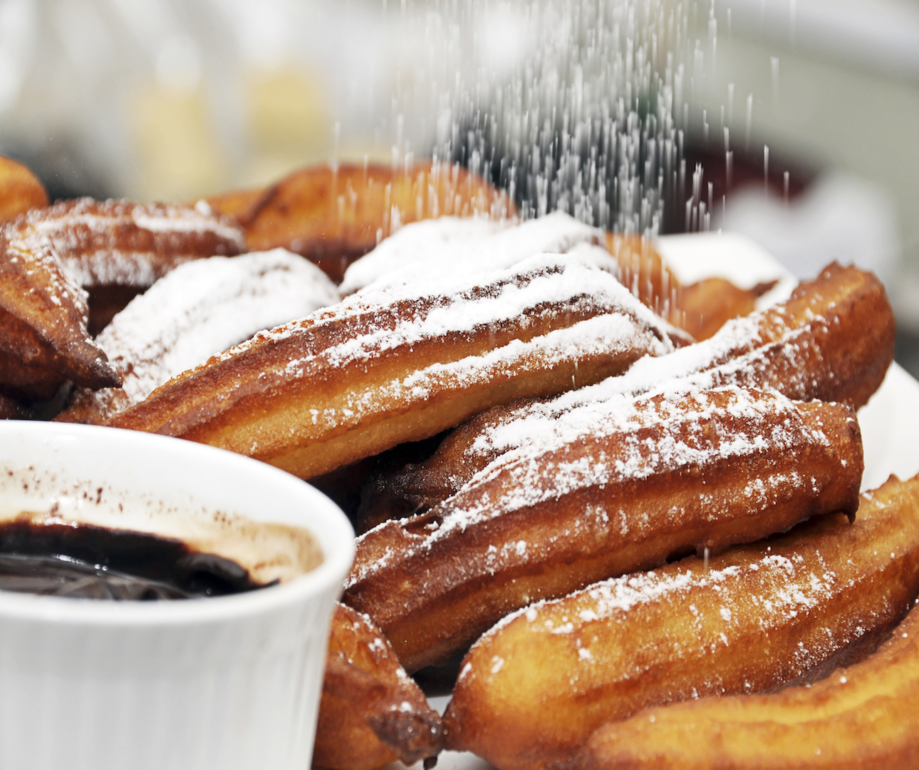 A row of churros lie while sugar rains down from above, coating them with a soft layer of white powder. A pot of dark sticky chocolate dip sits nearby.