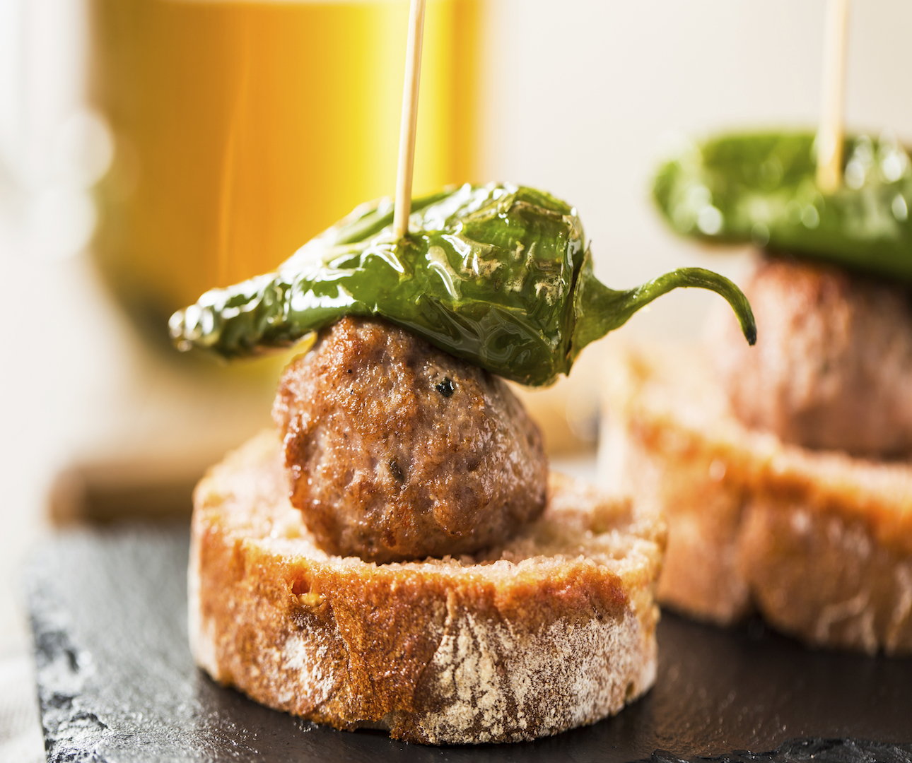 Closeup of a tapas-style dish of bread with meat and a pepper stacked on top of one another, held together by a wooden skewer