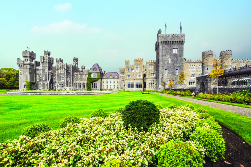 Grey brick currents rise against a bright blue sky with green lawns stretching down a driveway at Ashford Castle, ireland