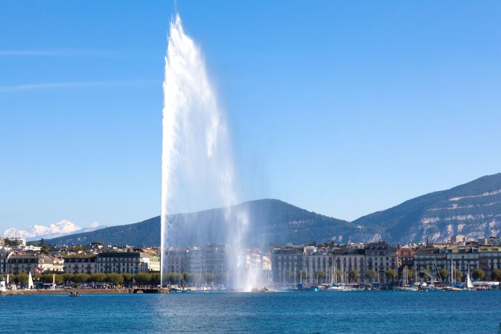 A high water jet on Lake Geneva Switzerland sprays water into the sky against the city backdrop and mountains.