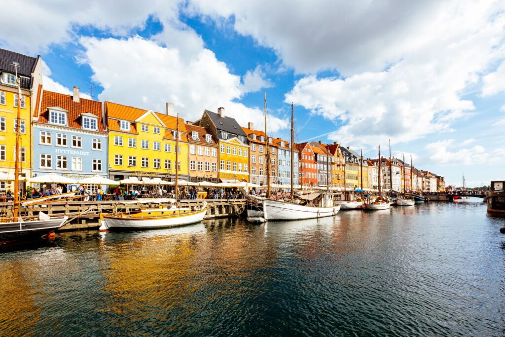 Colourful houses line the river in Copenhagen, Denmark, with boats and a blue sky with white clouds. 