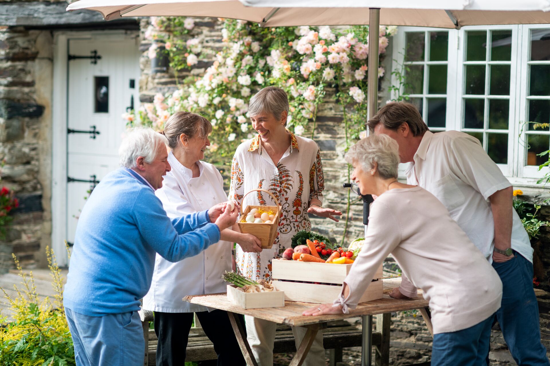 A group of senior people gather around an outdoor table leaden with fresh fruit, vegetables and eggs, in a. country garden with pink roses.