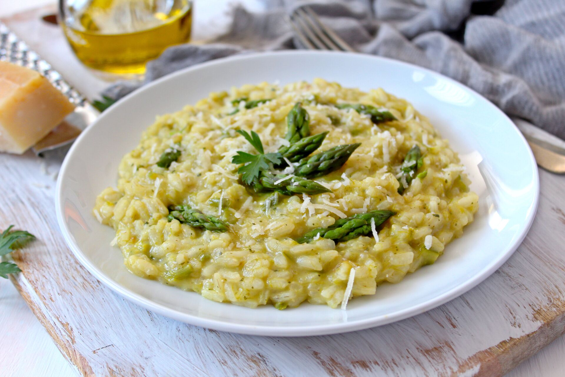 A white plate full of pale yellow risotto, with dark green herb leaves and slithers of cheese, is served on a wooden table in Italy.