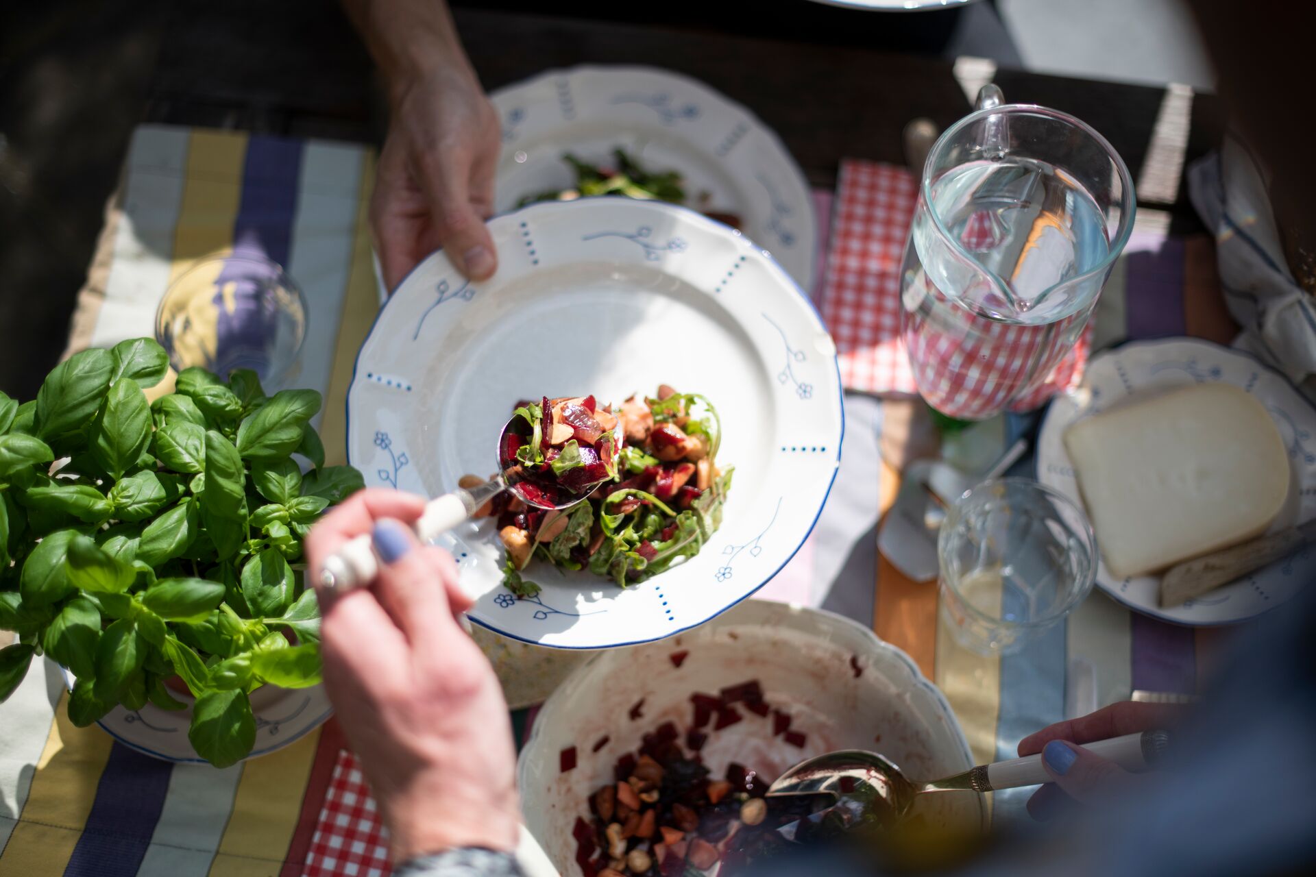 Aerial shot of a vegetable salad being served onto a white plate, with green herbs and cheese on the table alongside.