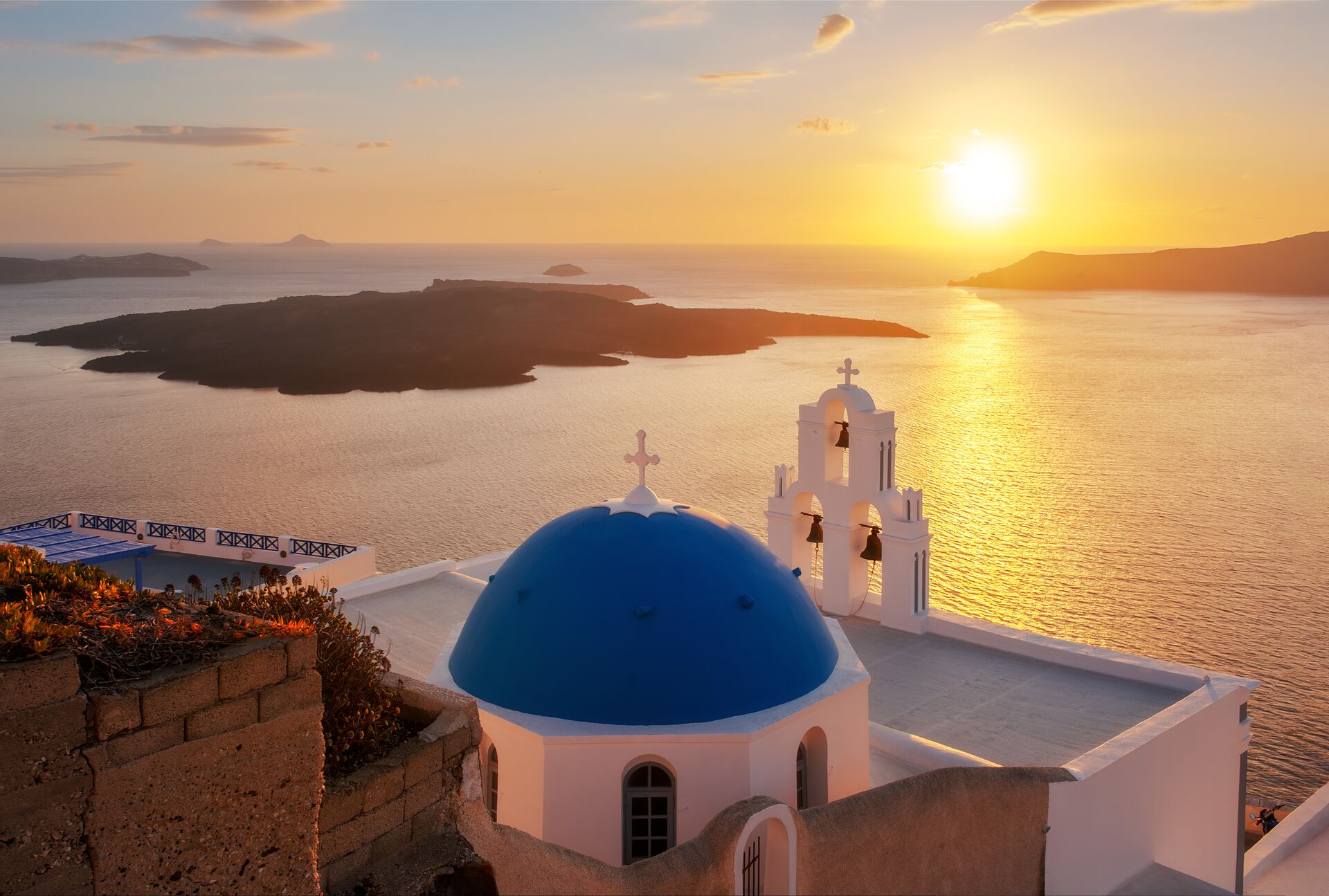 A White House with a bright blue domed roof looks out over the sea with the sun setting in th ebackgroundin Santorini, Greece.