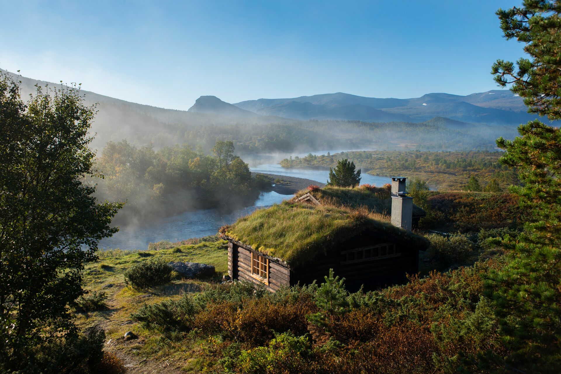 A timber cottage with a miss roof in a green fields beside a bright blue river.