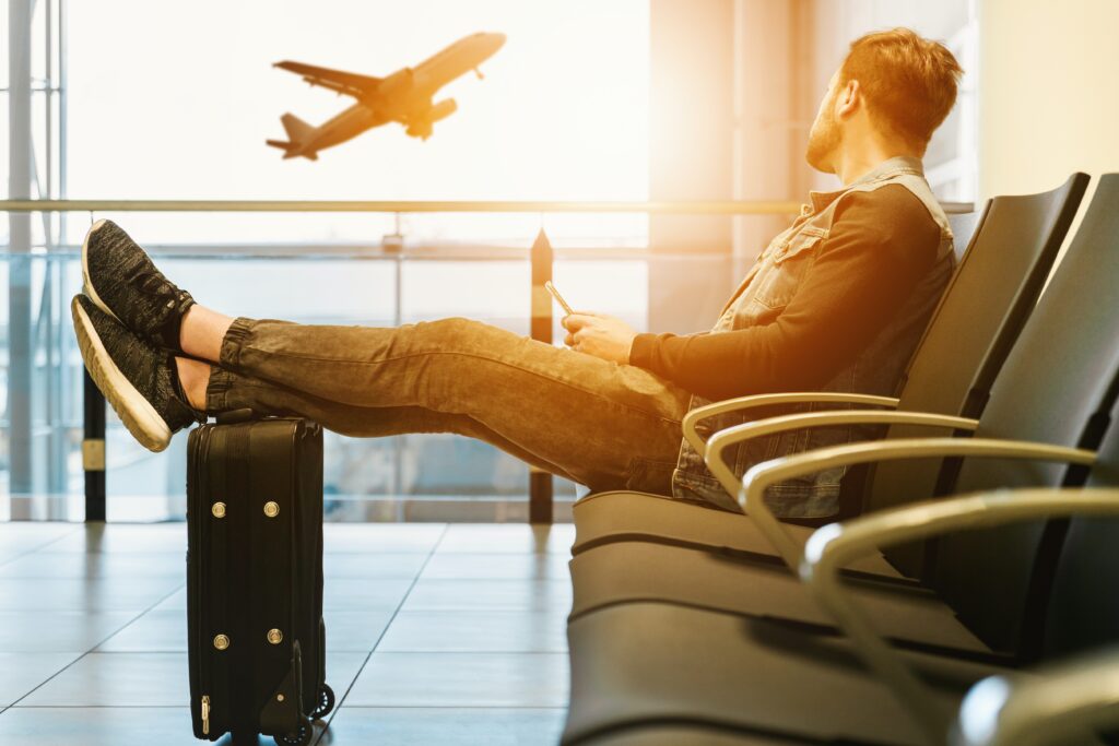 Image of man waiting at airport on a seat with his feel on his suitcase, with a plane taking off in the background