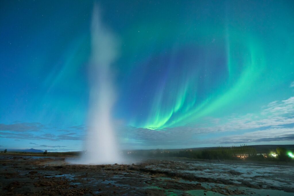 Green northern lights dance in a bright blue sky with a geyser in the foreground, in Iceland