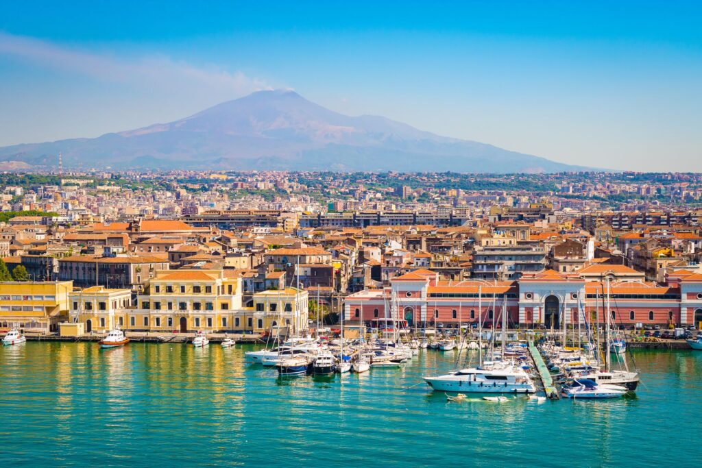 Image of fishing village in Sicily with bright waters in the foreground and colourful houses shining in the sun, a bright blue sky and Mount Etna in the background