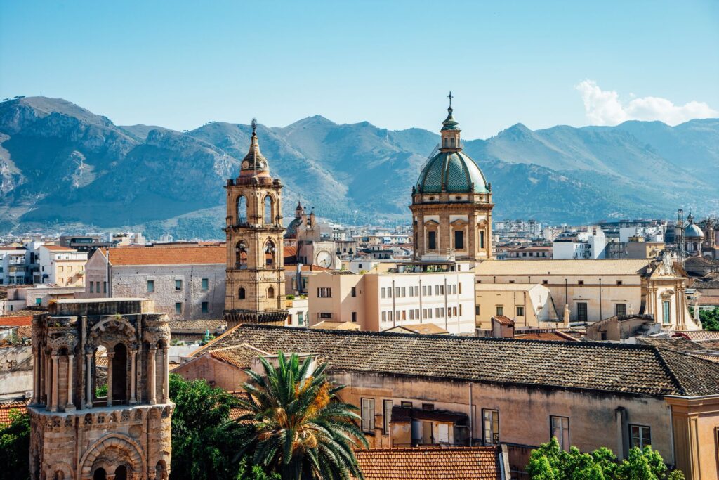 The spires of Palermo stand out above buildings, with mountain and a hazy blue sky in the background.
