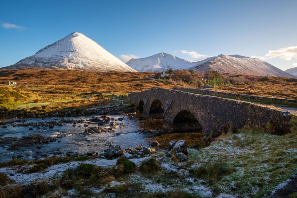 A stone bridge leads across a river, with snow capped mountains in the background