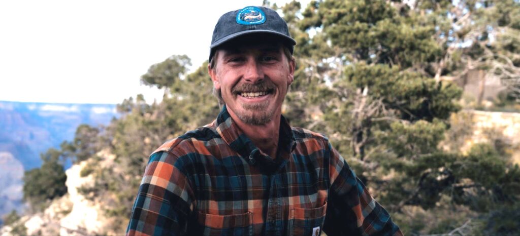 A man in a plaid shirt is smiling in front of a guide to the Grand Canyon.
