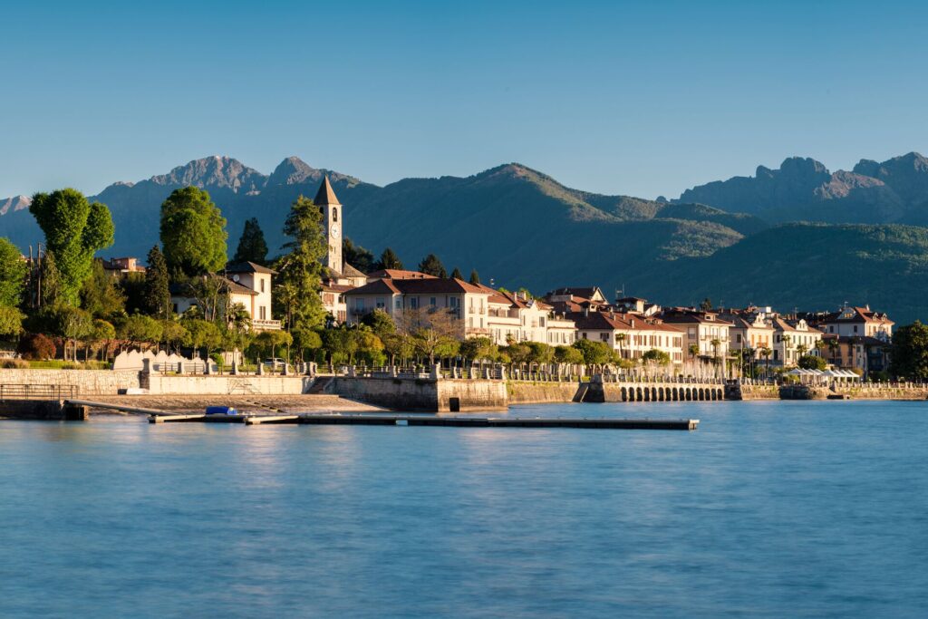 Cream coloured houses with red roofs sit on the edge of Lake Maggiore with blue waters and mountains in the background.