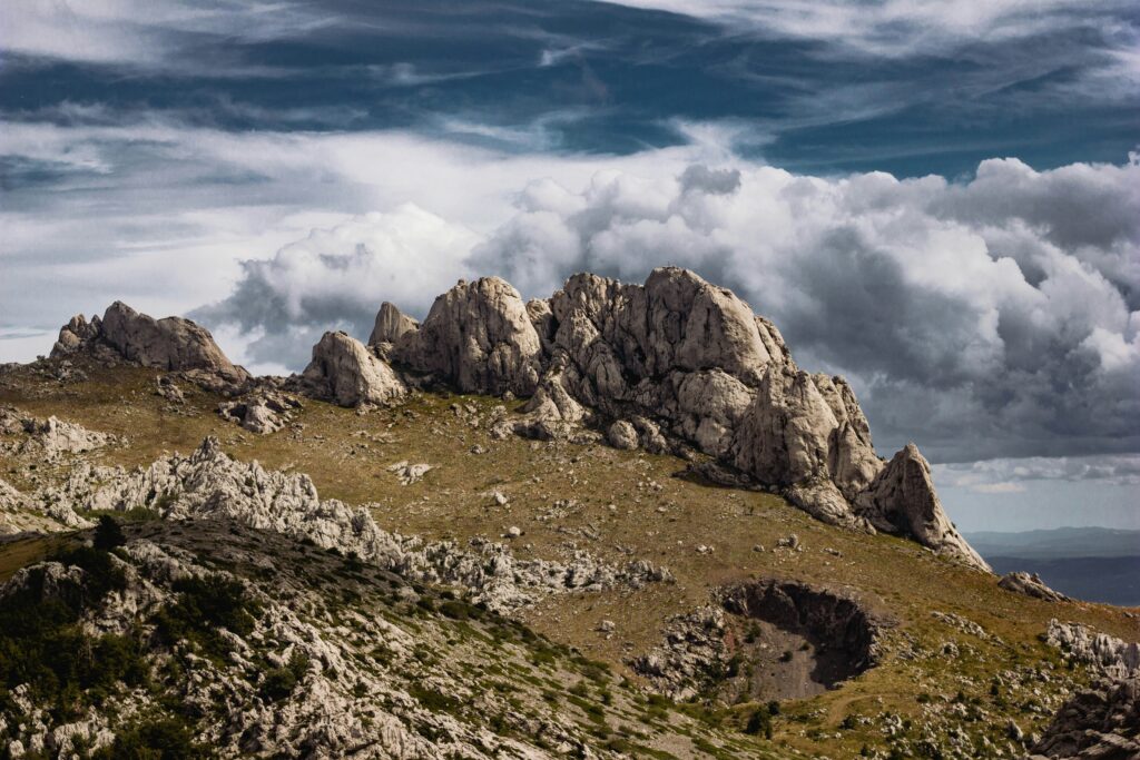 Craggy mountains with rick formations, with billowing clouds and a dramatic grey sky in the background.