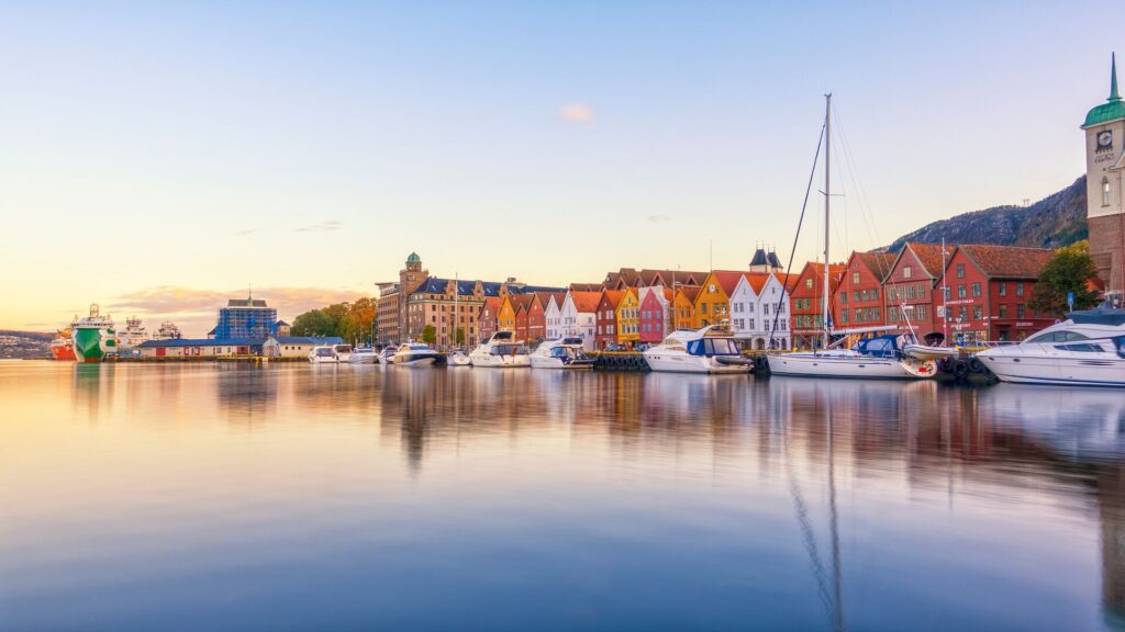 Red, white and orange houses of Bergen, Norway sit colourfully along the water's edge, with boats on the gleaming water.