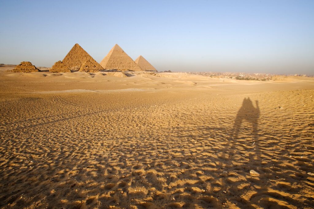 The Pyramids of Giza in Egypt in the background and a foreground of sand with the shadow of a camel.