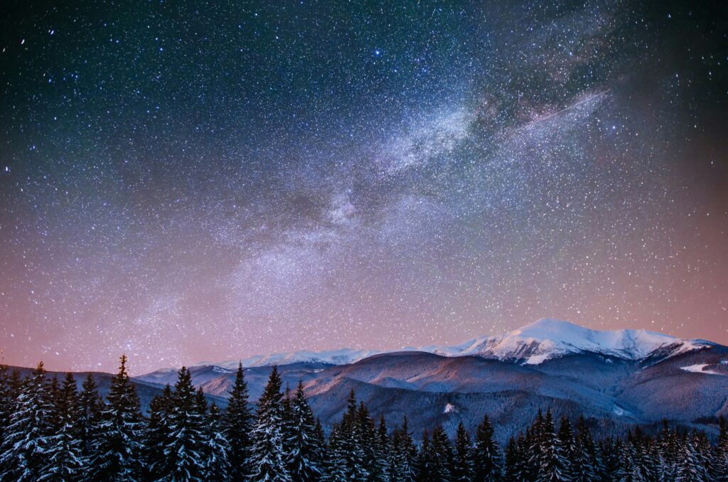 The Milky Way in the night sky, over a forrest and mountains.