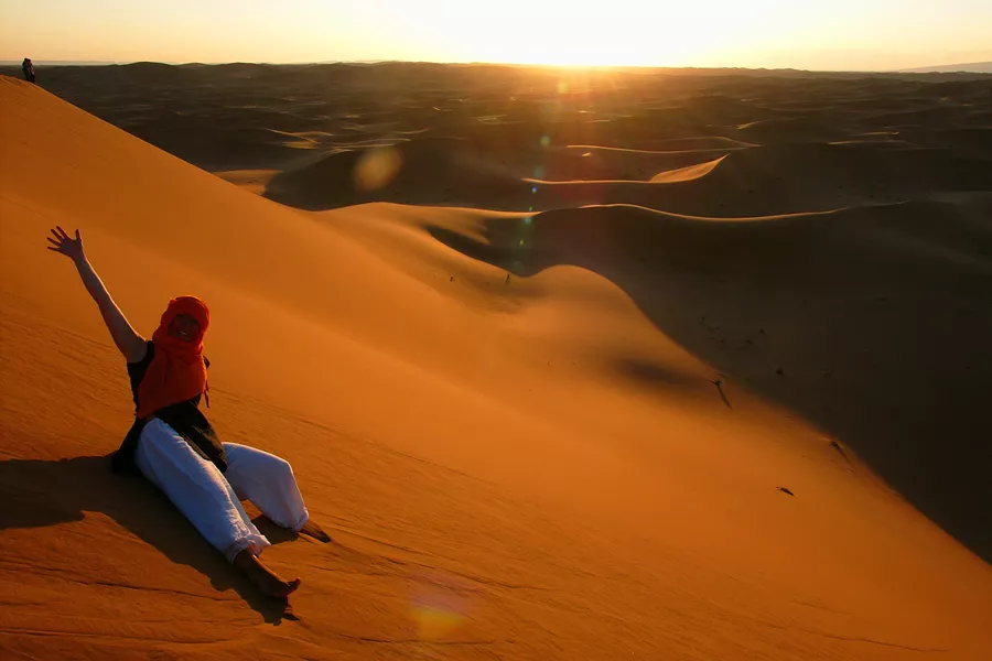 Smiling Young Woman With Arm Raised Sitting On Sand Dune During Sunny Day 674746141