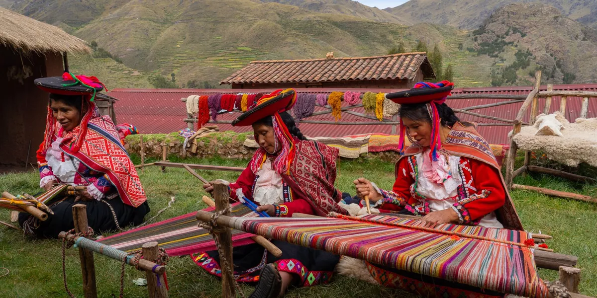 Three local female weavers in colourful traditional local dress including festooned hats, weaving colourful alpaca wool on the ground