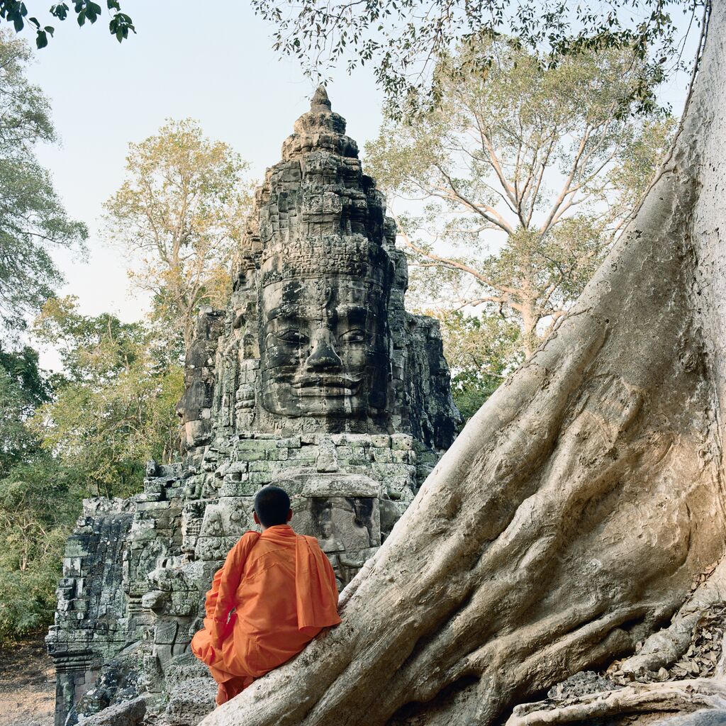 Buddhist monk sitting on tree roots at Angkor Wat, infront of ancient stone carving in Cambodia