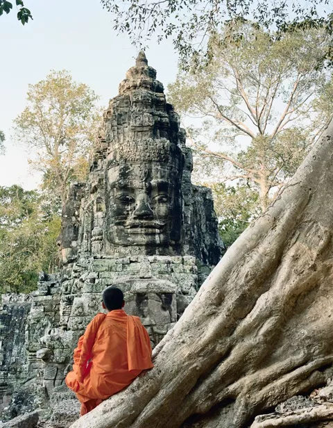 Buddhist monk sitting on tree roots at Angkor Wat, infront of ancient stone carving in Cambodia