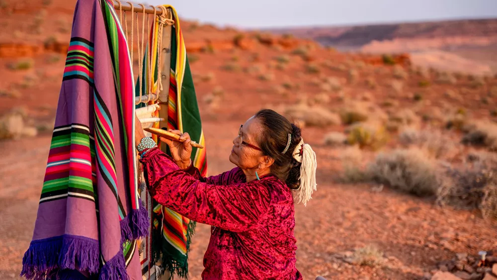 Elderly Navajo Woman Weaving A Traditional Blanket Or Rug On An Authentic Native American Loom In The Desert At Dusk Near The Monument Valley