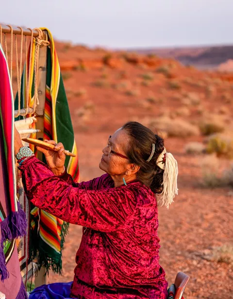 Large Elderly Navajo Woman Weaving A Traditional Blanket Or Rug On An Authentic Native American Loom In The Desert At Dusk Near The Monument Valley Tribal P 1278436213