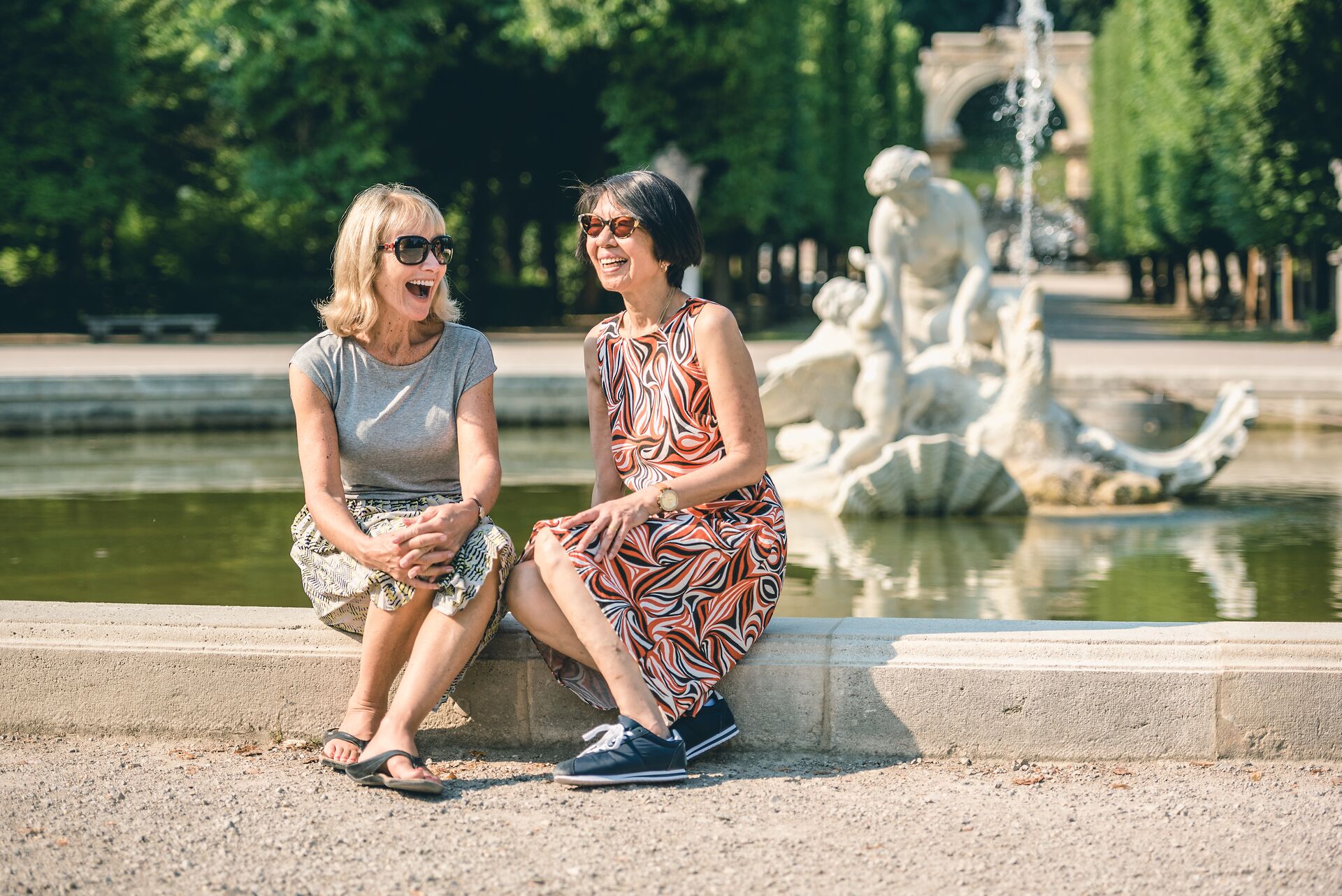 2 woman laughing while sitting next to a fountain in Schonbrunn Palace Gardens in Vienna, Austria