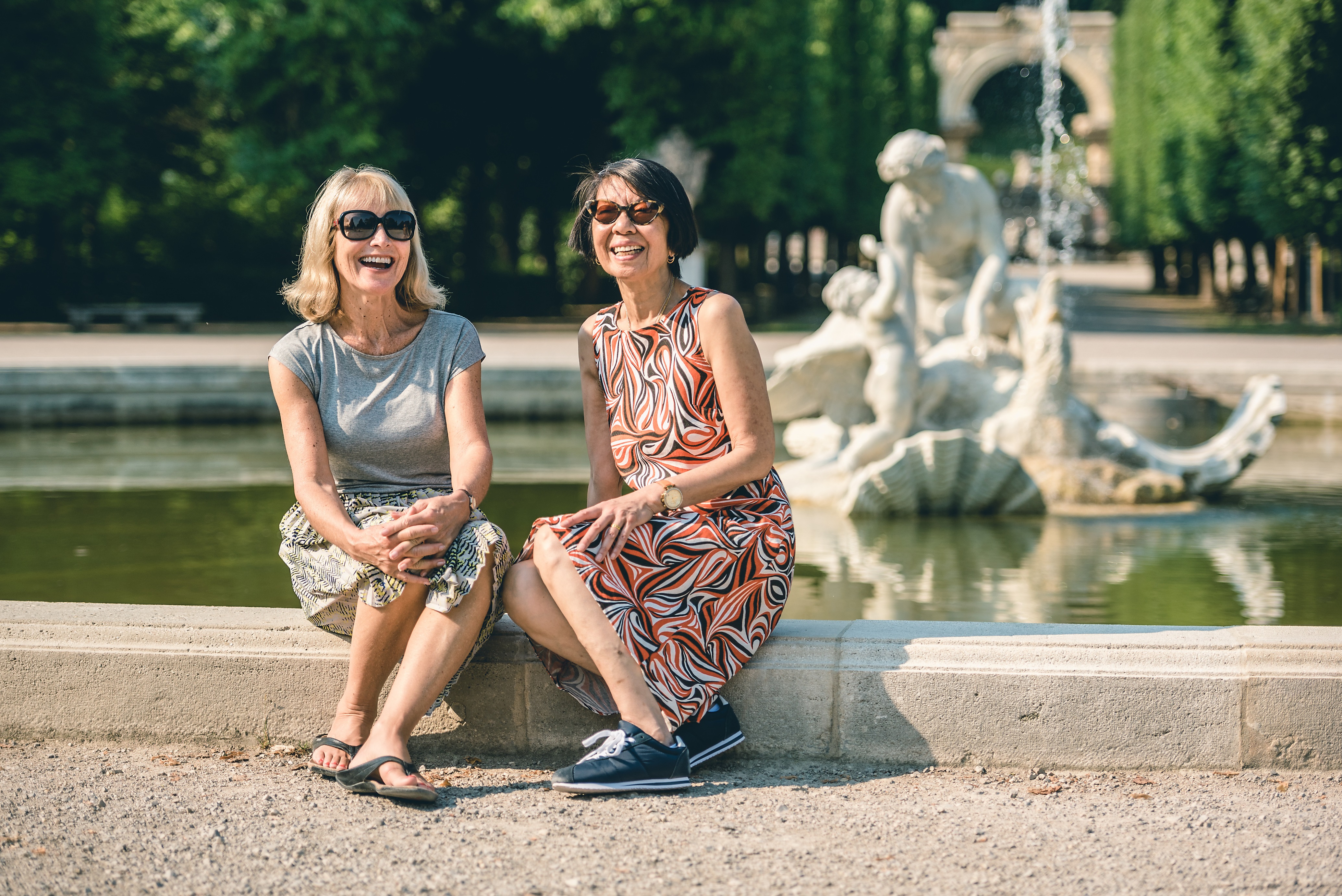 Two women sitting on a fountain and smiling.
