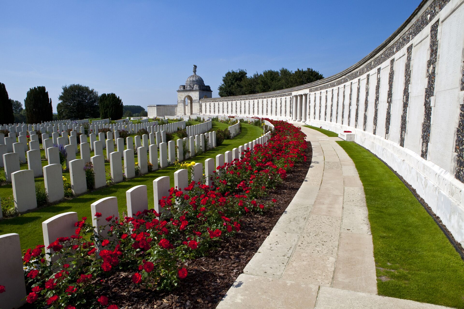 A large cemetery in Ypres Belgium with semi circular rows of white gravestones, rows of red flowers and a white memorial wall.