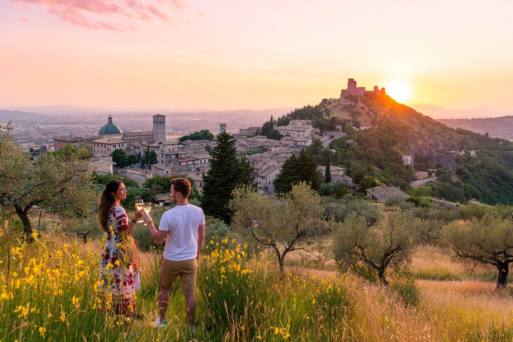 A couple enjoying a beautiful view of Italian hillside villages stretching out into the horizon at sunset. They hold glasses of wine and are surrounded by spring flowers