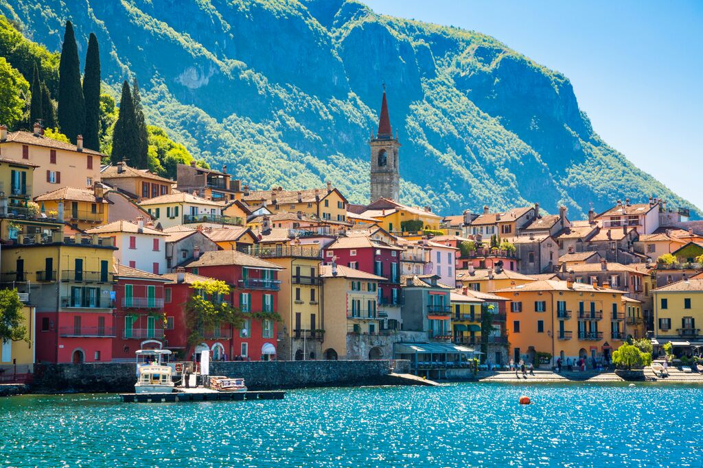 A beautiful Italian village glowing in the sun, multi-coloured houses look out over a glittering body of water. A grass-covered mountain makes up the background