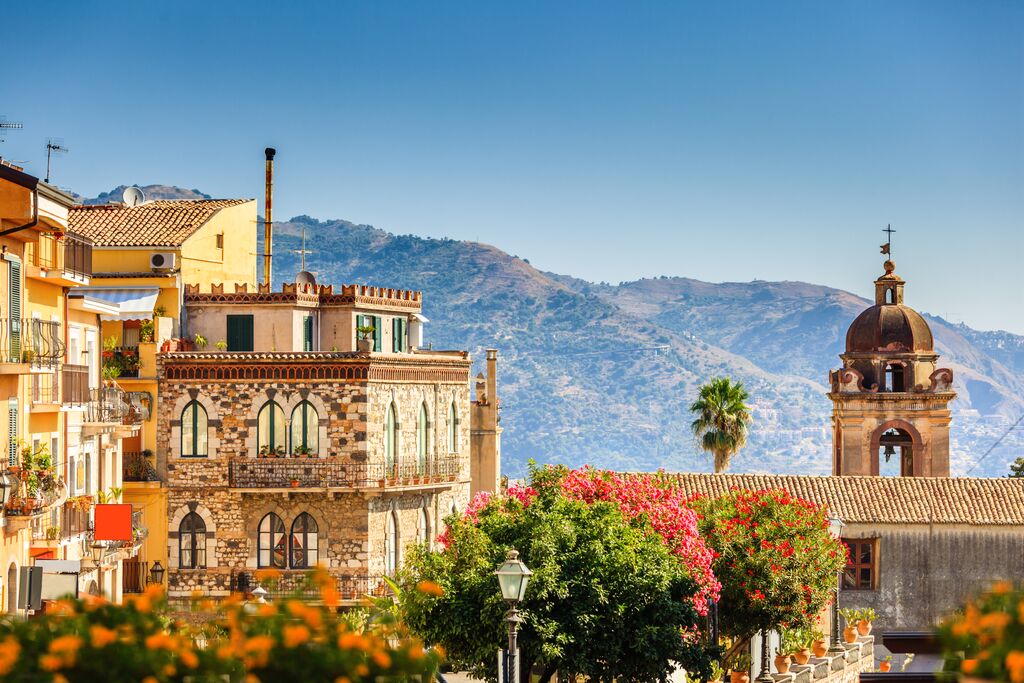 A view of Taormina buildings backdropped by towering hilltops. In the foreground, blooming trees and bushes with red and orange flowers.