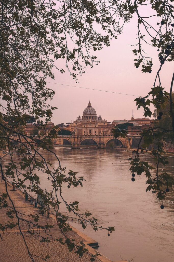 A classic Italian building framed by tree leaves, sitting in front of a body of water. The sky is pink in the may sunrise