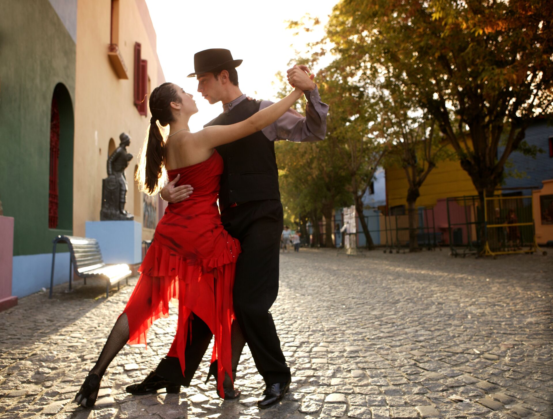 Image of young couple dancing tango in a cobbled street. The lady is wearing a bright red dress and the man is in black, with the sun, houses and trees behind them