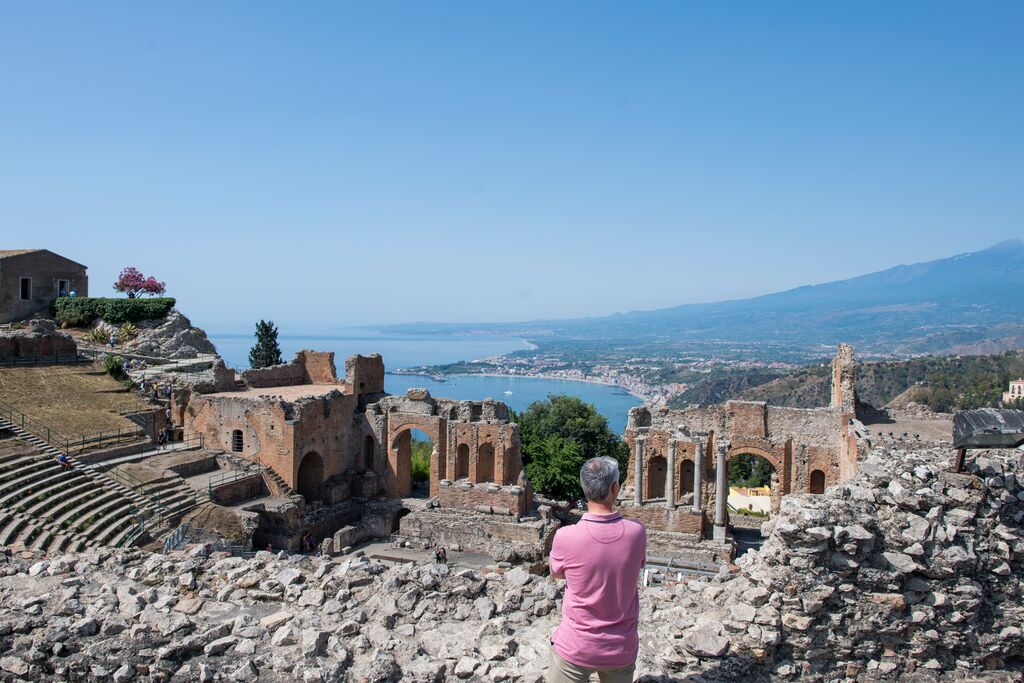 Old ruins in Sicily, backdropped by gorgeous mediterranean coastline. A man in a pink top looks out into the distance.