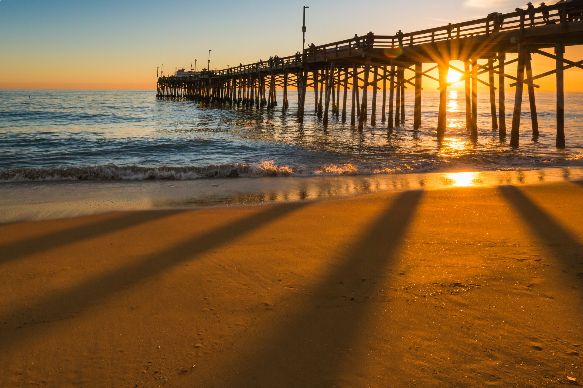 Image of sunset behind a pier, with the rays casting warm light and shadows across the ocean and sand in the foreground