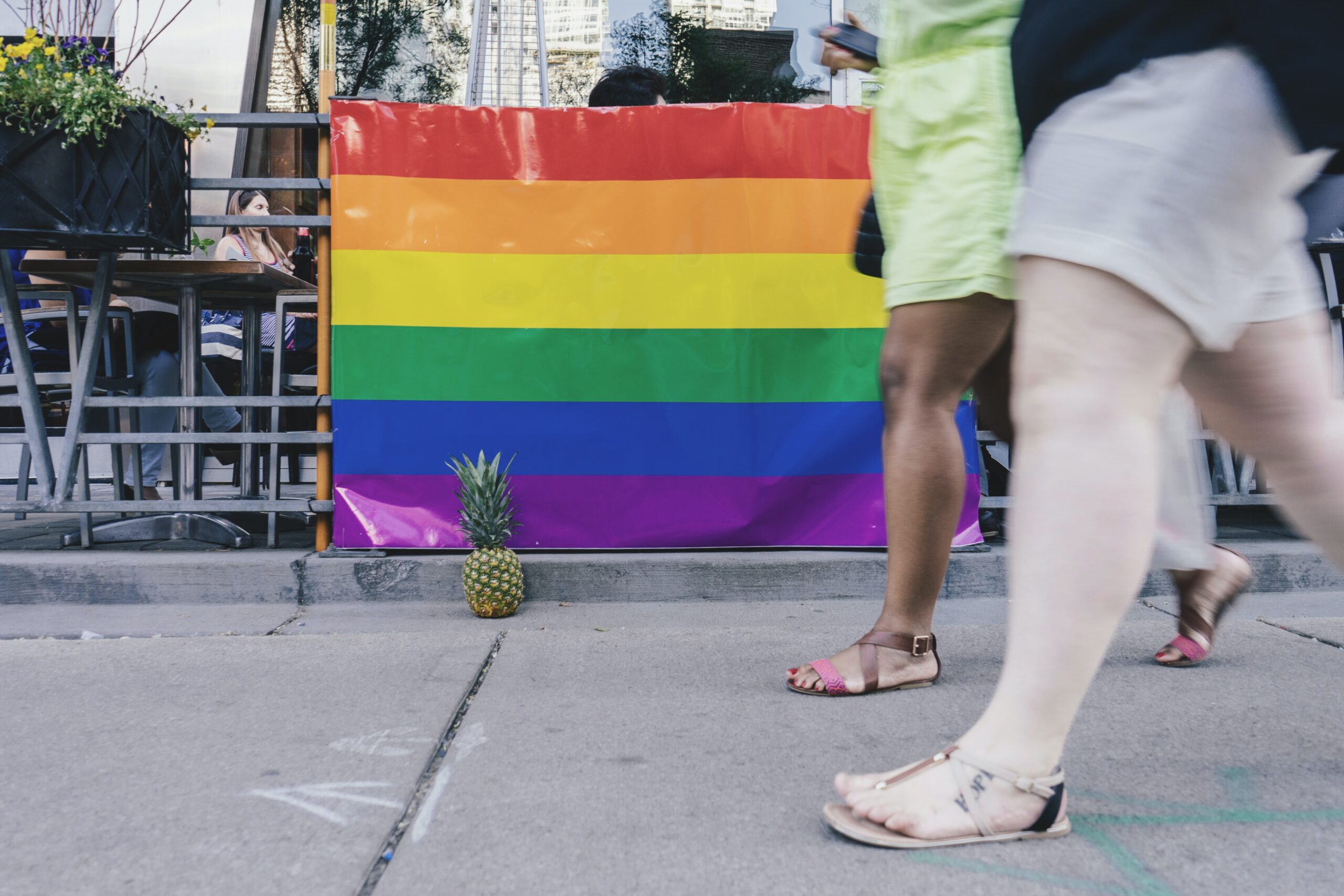Ground level image of legs walking in front of a bar displaying a rainbow pride flag, showing it is open to inclusive travel