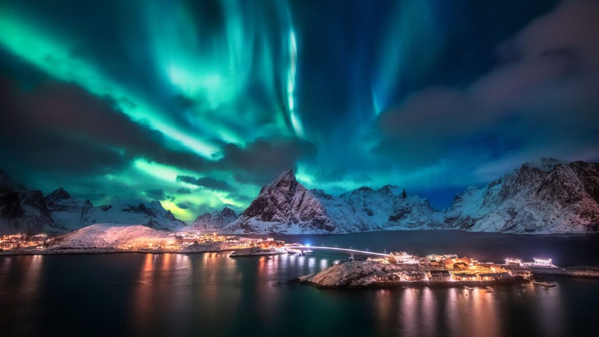 Bright green and blue northern lights against the night sky with water in the foreground and mountains in the back