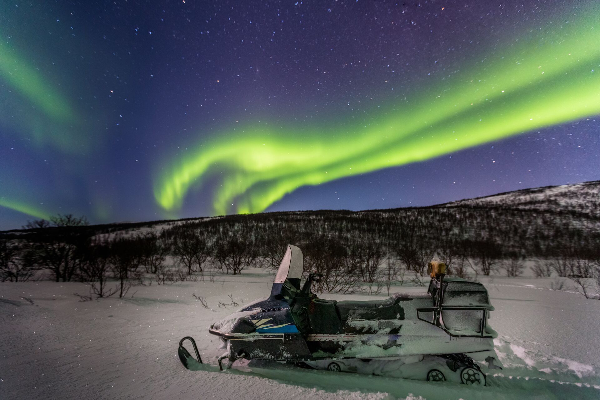 three green northern lights lines track across the purple night sky, with show and a snowmobile in the foreground