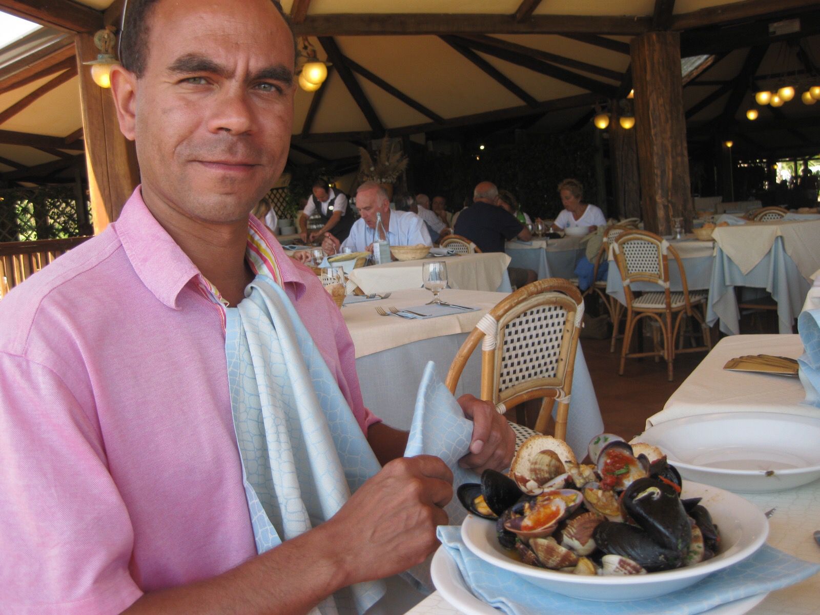 Marks, an expert in Sicilian cuisine is pictured seated at a local restaurant, wearing a pink shirt and a napkin enjoying a bowl fo seafood