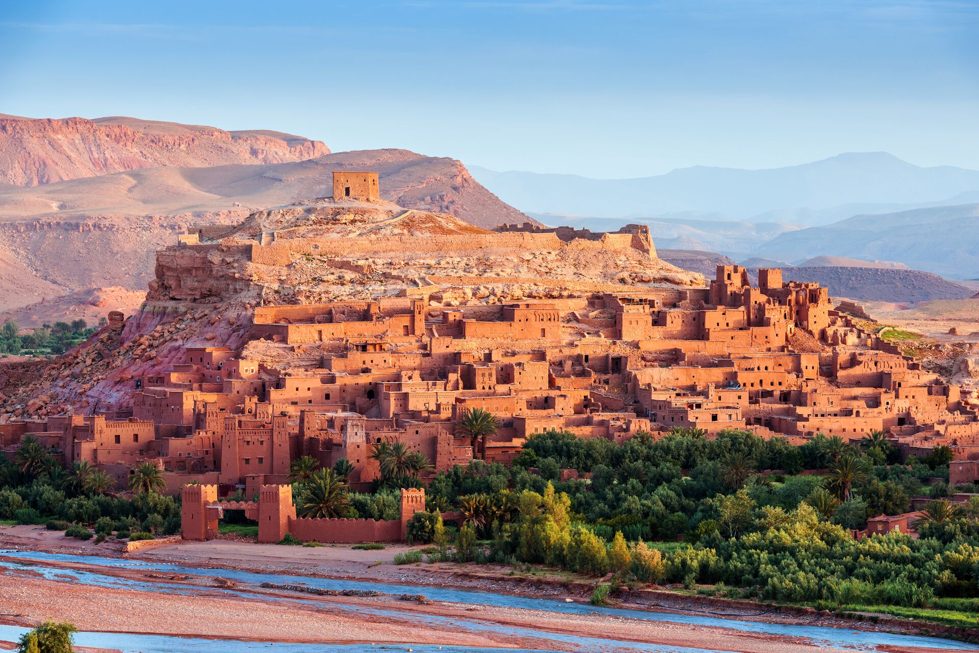 Red stone houses of the Ait Ben Haddou Kasbah sit in a red sand valley with a bright blue sky and red mountains in the background