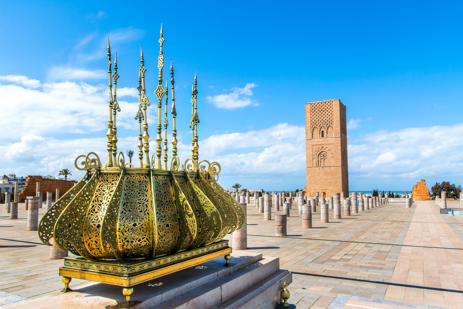 Green and gold ornate Mausoleum of King Mohammad IV shines in the Moroccan sun, situated on a light brick courtyard with a tower in the background and a bright blue sky
