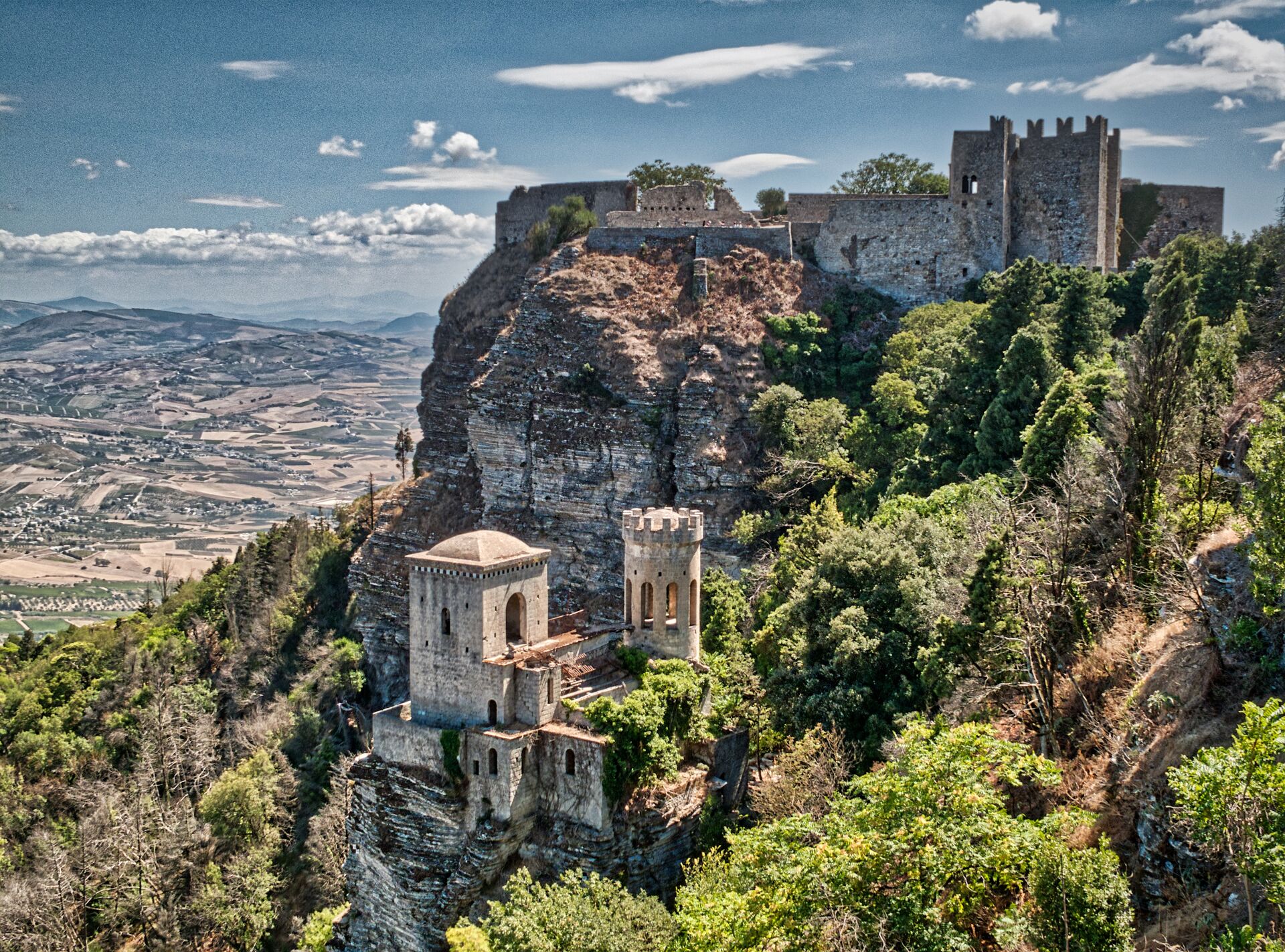 Erice castle in Sicily is perched high in a montuanside surrounded by green trees with the fields stretching out below and behind
