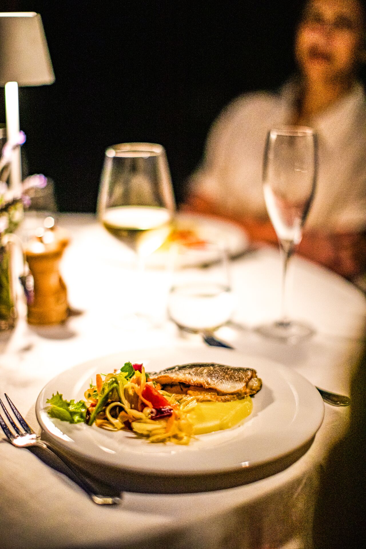 an elegant plate of fish with vegetables shows farm-to-table fine dining in sicily, on a white table cloth with a glass of white wine