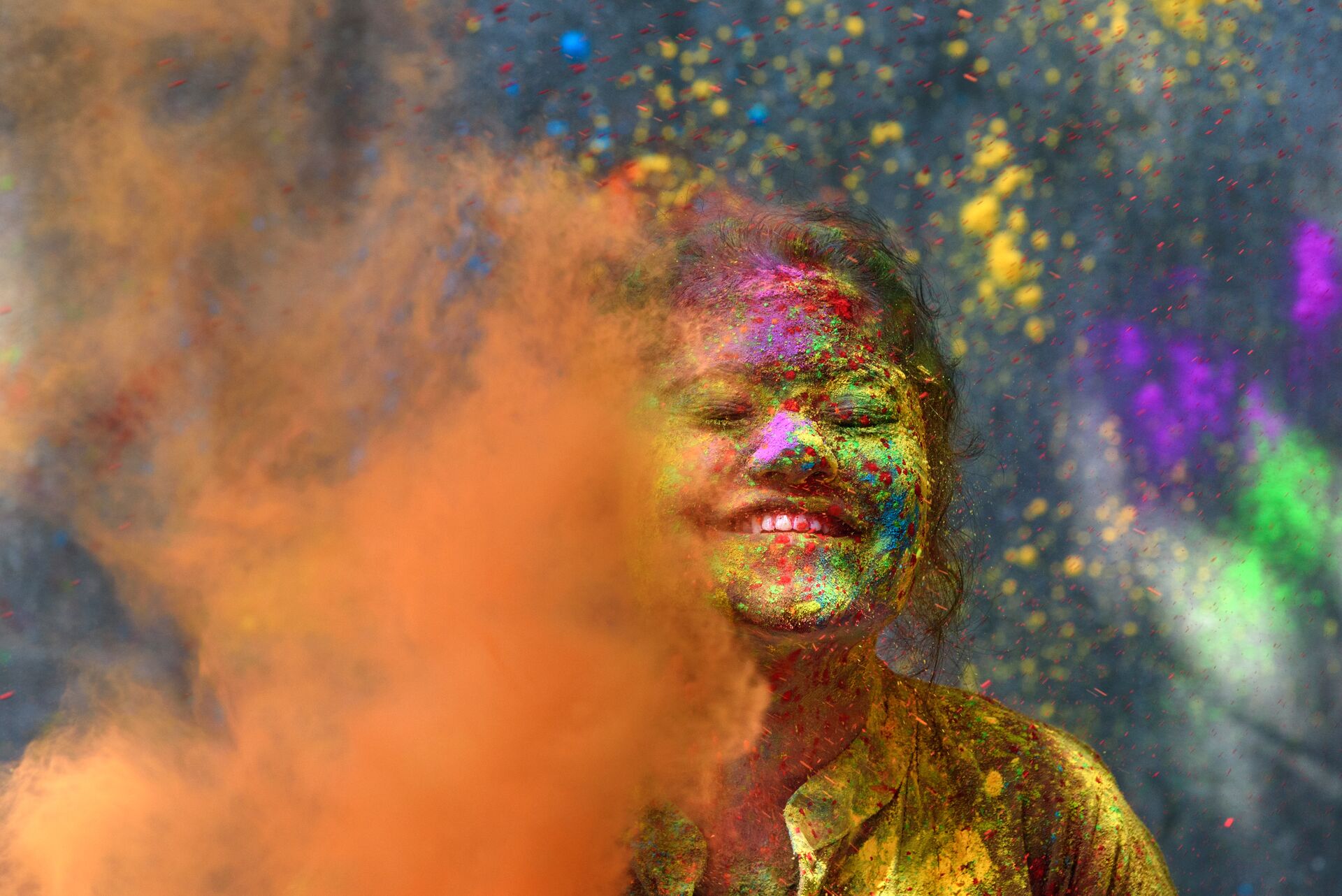 close up of young girl with colourful powder all over he face and more being thrown in front of the picture, a mix of organic, pink, yellow and green during holi spring festival