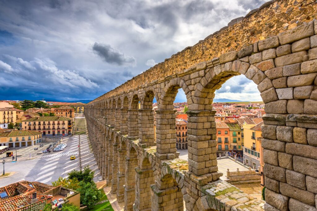 Segovia’s stone aqueduct is show going away from the camera, with town buildings in the background and a cloudy sky