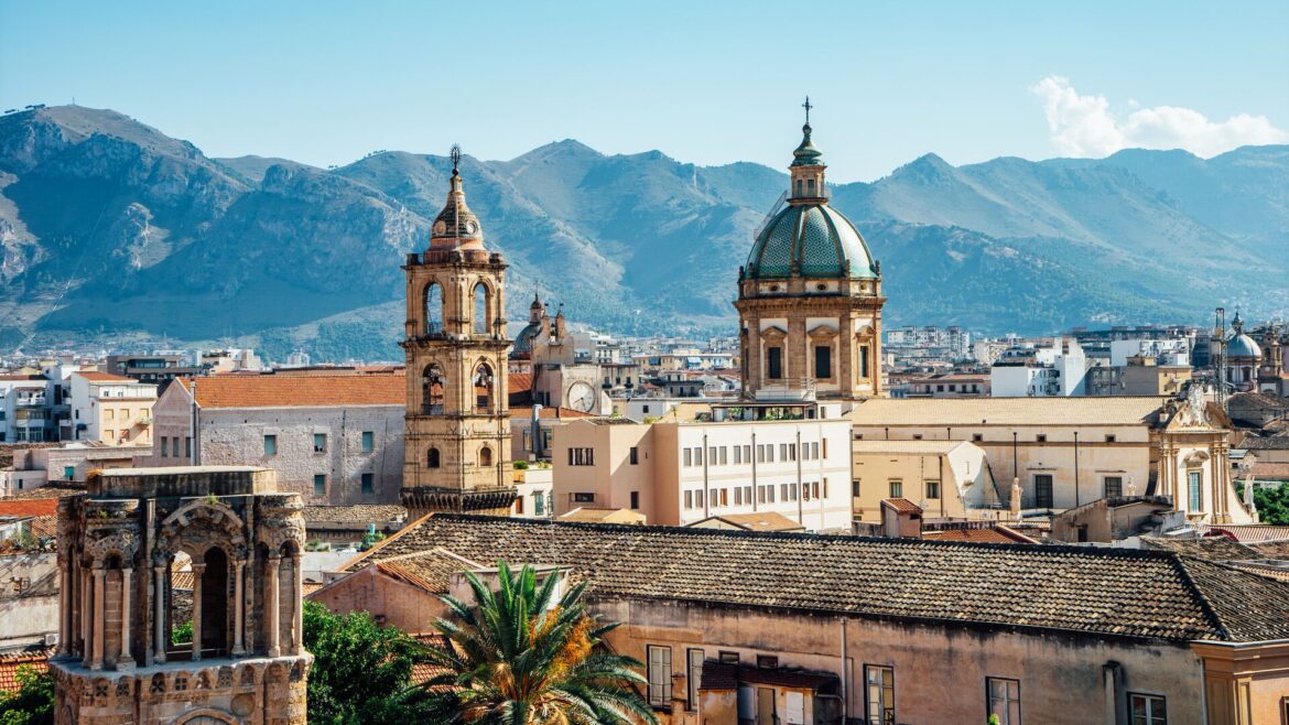 The spires of Palermo stand out above buildings, with mountain and a hazy blue sky in the background.