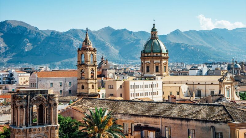 The spires of Palermo stand out above buildings, with mountain and a hazy blue sky in the background.