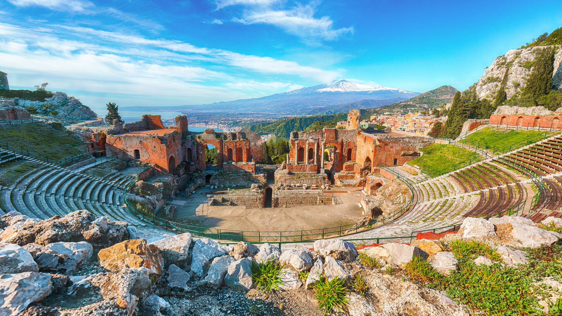 Image of Ancient Greek amphitheatre in Taormina, a redish stone stage with seating flowing up the sides, with spectacular views across the hillsides and a bright blue sky